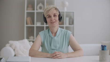 charming-middle-aged-woman-with-headphones-is-greeting-by-videocall-looking-at-camera-sitting-in-room-medium-portrait